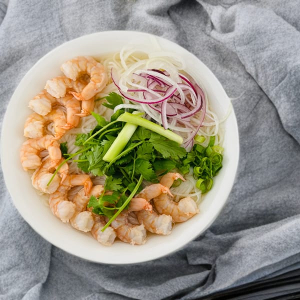 A white bowl of Chef's Phở & Grill shrimp pho with noodles, cilantro, green onions, and red onions.