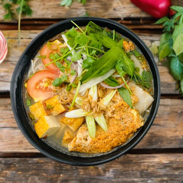 Overhead view of a Vietnamese noodle soup with tofu, tomatoes, and fresh herbs in a black bowl.